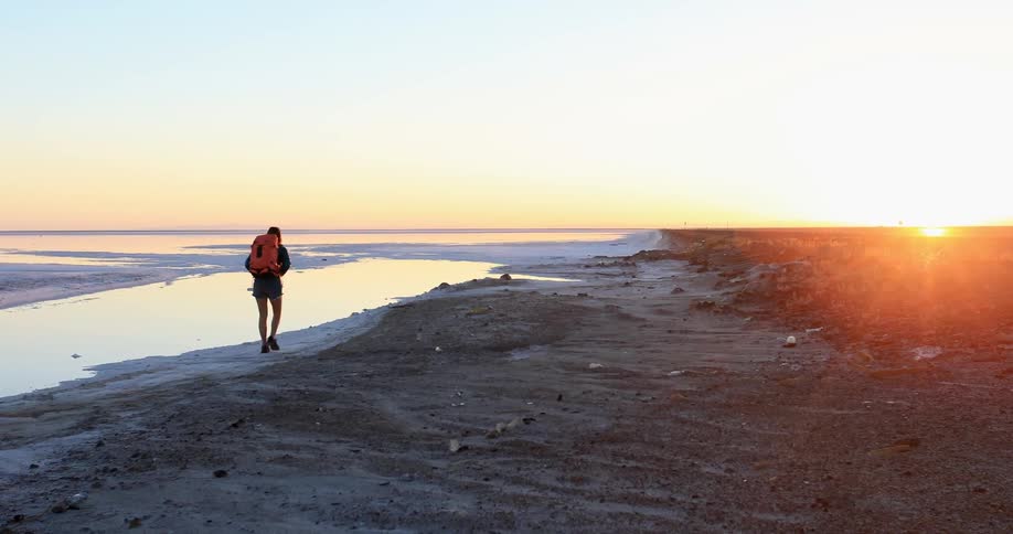 A woman wearing a backpack walks briskly along an empty beach at sunset