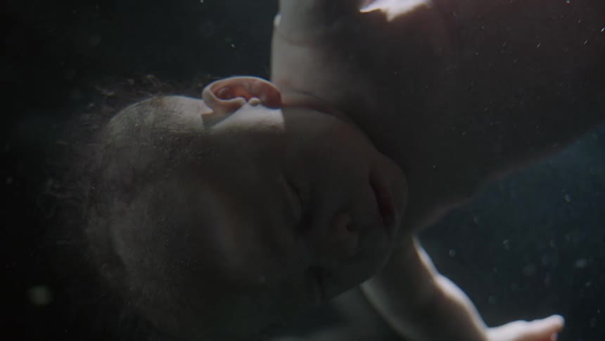 A baby floats in liquid with eyes closed and lights shining and then going dark