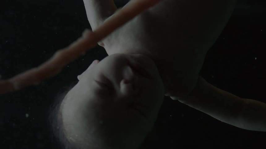 A baby floats upside down in liquid with a cord showing with strobes of light and then darkness