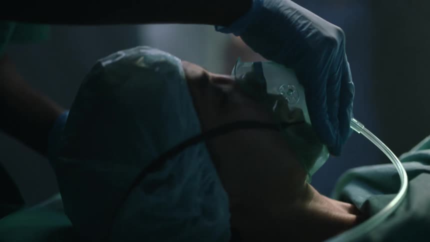 A woman lying on a gurney in an operating has an oxygen mask placed on her face by medical staff