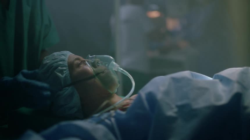 A doctor places an oxygen mask over a womans mouth and nose as she turns her head to look at doctors working on her baby
