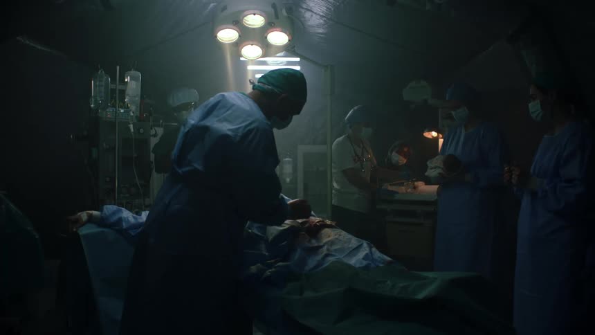 A doctor pats the bloody sheet over a woman who has just given birth via c section as the nurse carries the baby over to mom