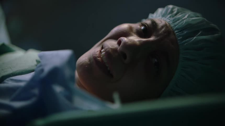 A woman is lying on a gurney wearing a sterile hat nervously smiling