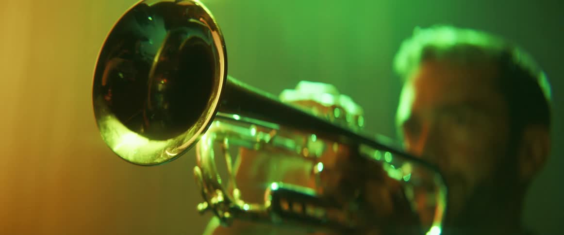 A man raises a trumpet to his lips while standing indoors under colored lighting