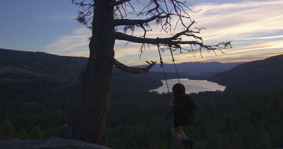 Woman wearing a hat swings on a tree swing in front of a lake at sunset
