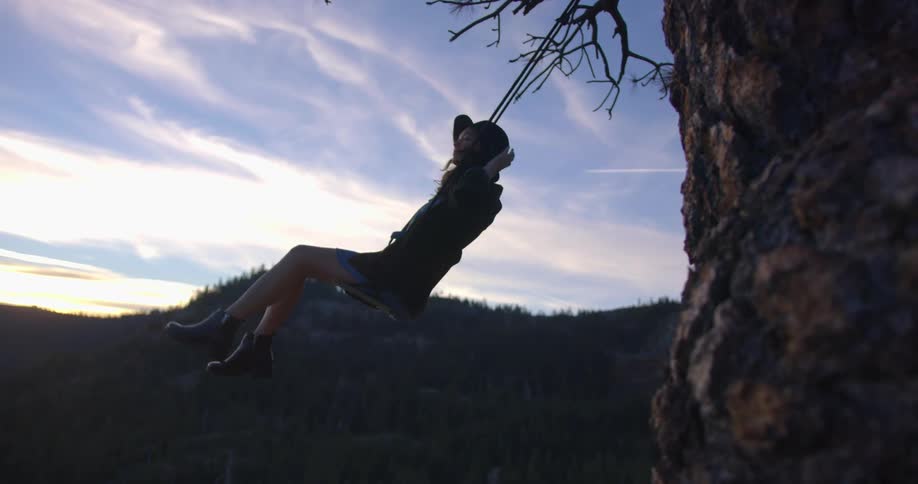 A young woman swings back and forth in a tree swing on a mountain slope on a cloudy evening