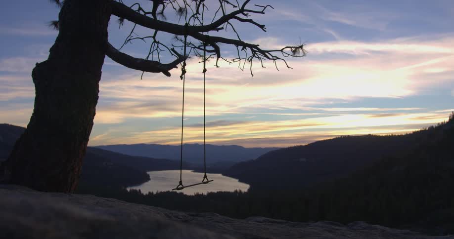 A tree swing on a rocky mountain slope overlooking a forested valley containing a lake