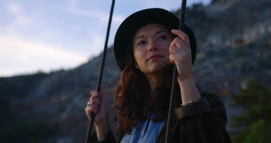 A young woman sits on a spring outdoors in front of a hill at dusk