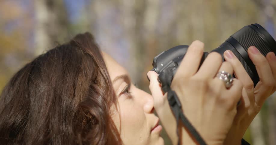 A young female photographer looks up at the trees of a woodland before raising her camera to her eye to take a photograph