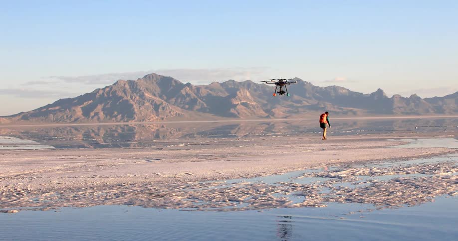 A drone flies behind a man as he walks across a large flat basin