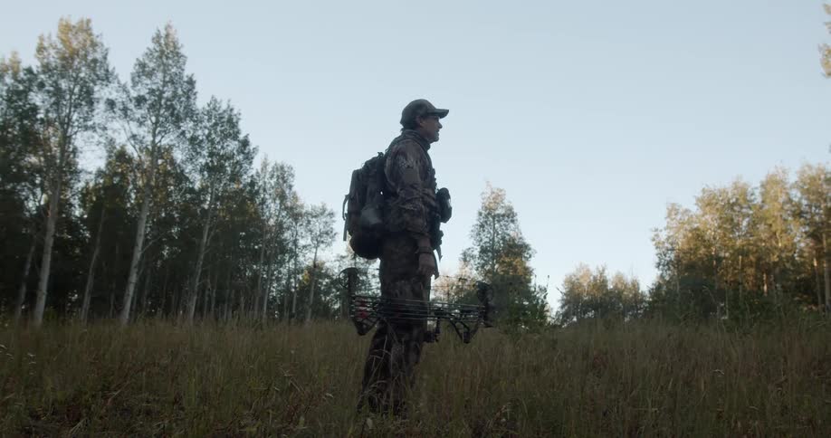Man holds hunting bow while standing in field near woods