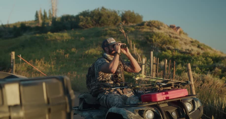 A man wearing camouflage sitting in an all terrain vehicle uses binoculars at sunset