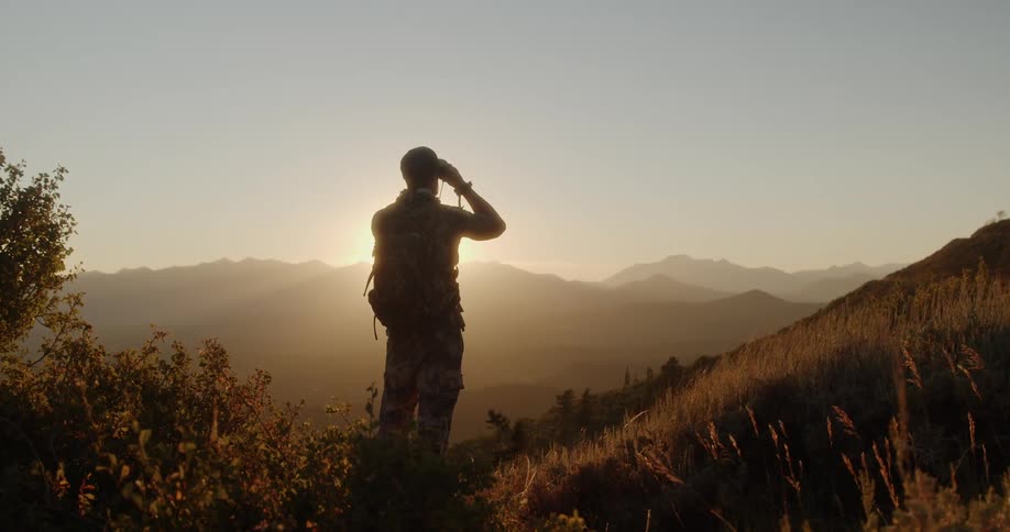 A man in camouflage uses binoculars to look across the landscape as he stands on a hill
