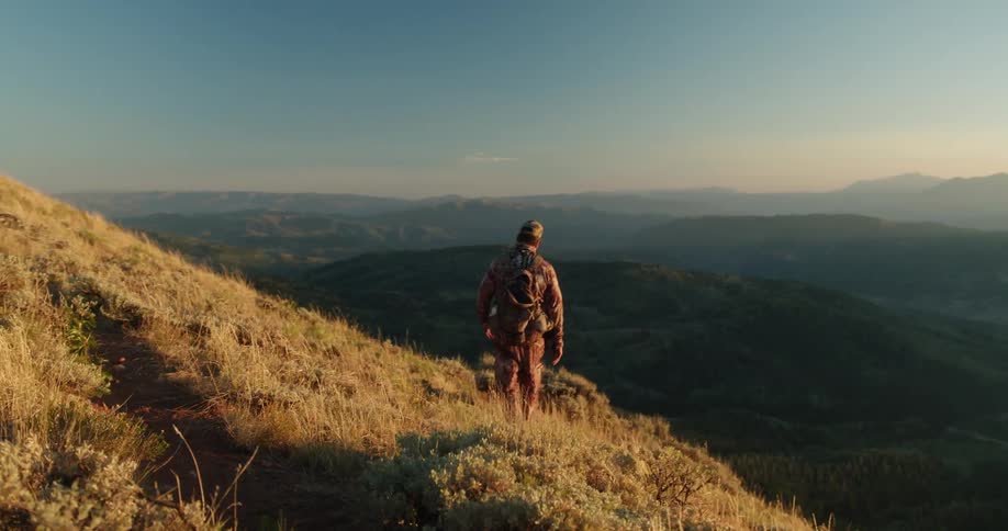 Man with a cap and backpack walks downhill with mountains in the background at sunset