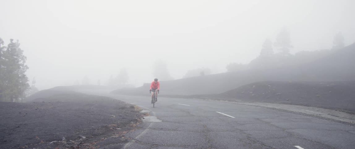 A cyclist struggles up hill along the curve of a remote mountain road through dense fog