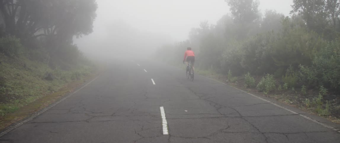 A man is riding a bike through the fog on an asphalt road
