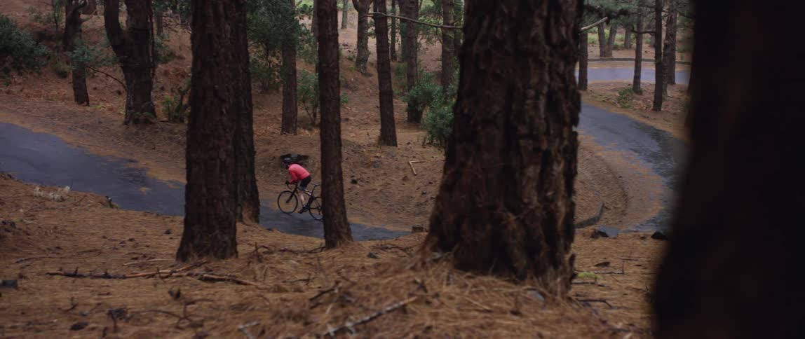 wide shot of a cyclist pedaling up hill on a winding forest road trail while its snowing