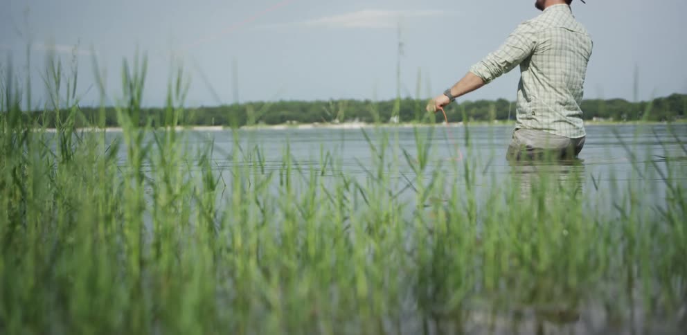 A fisherman casts his fishing line while standing hip deep in the waters of a lake during the day