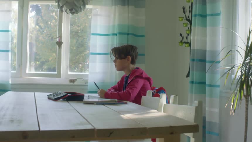 A boy writes on paper while sitting at a table in a home during the day