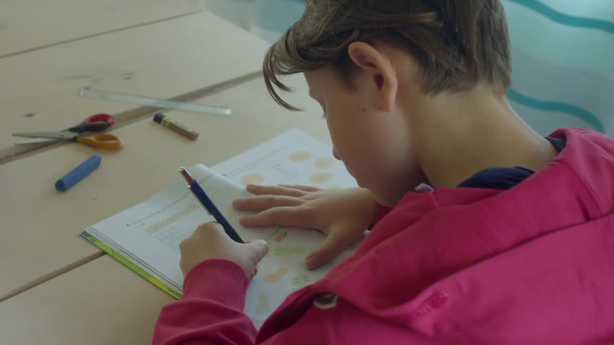 A boy writes in a workbook while sitting at his kitchen table with school supplies