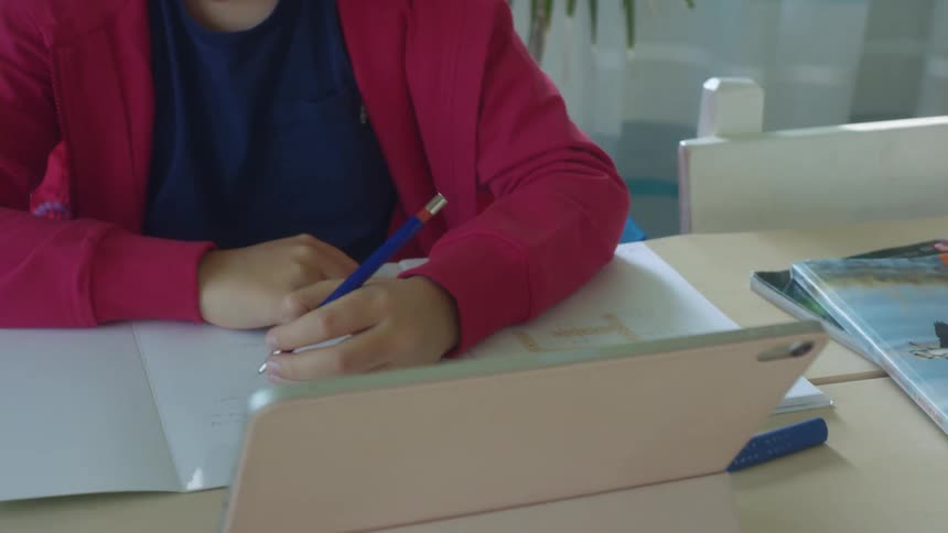 A child writes on paper while sitting at a table with a tablet on it as well as other books