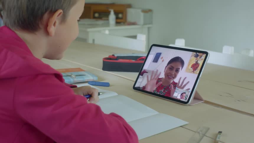 A student waves at his teacher on a tablet while sitting at his kitchen table