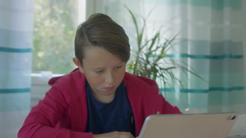 A boy sits at a table with a tablet writing on a piece of paper