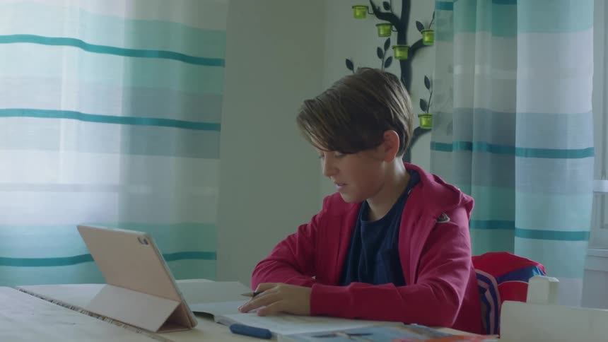 A boy sitting at a table with a notebook and textbook lying before him as he speaks to a tablet resting on a tablet stand on the tabletop