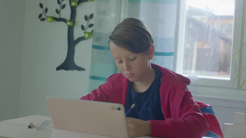 Boy attends classes via computer sitting near window showing nearby houses