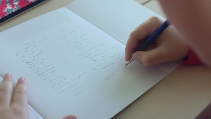 A student writes in a notebook with a tablet propped on the table in front