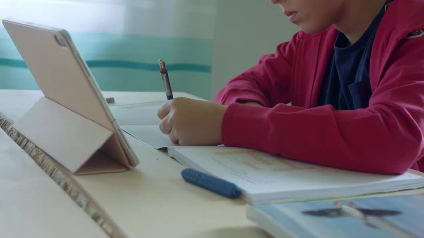 A boy write on paper doing homework at a table at home with a laptop in front of him