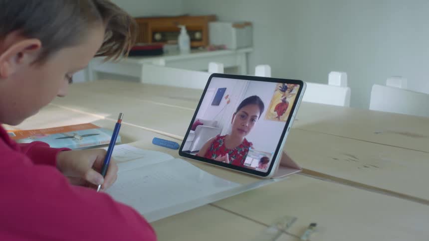 A young boy at home watches his teacher on a tablet and does school work