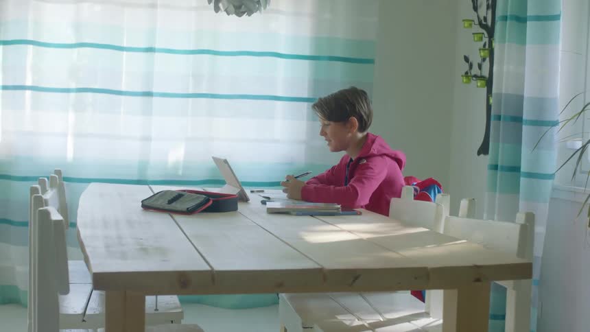A girl in a room sitting on a chair at a table in front of a tablet