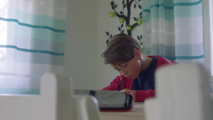 A boy sits at a home table writing with a pencil