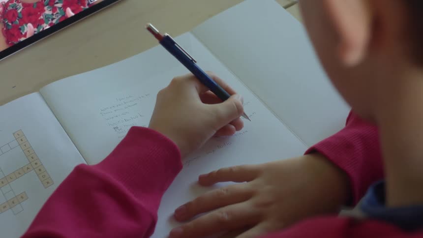 A boy sits at a desk writing on paper with a screen sitting in front of him