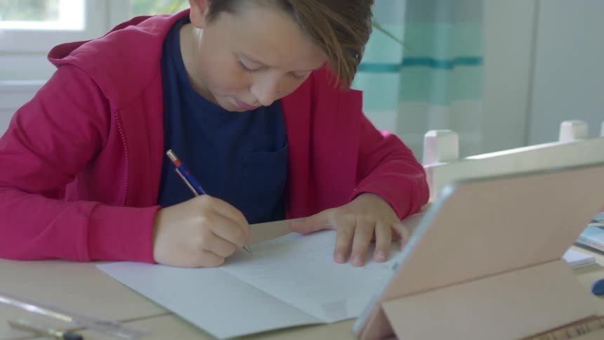 A boy sits at a large home table with a tablet having a video call and writing on a piece of paper