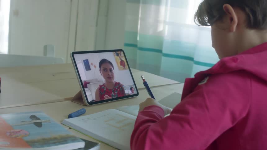 A boy sits at a table doing homework with an adult on a tablet sitting on the table