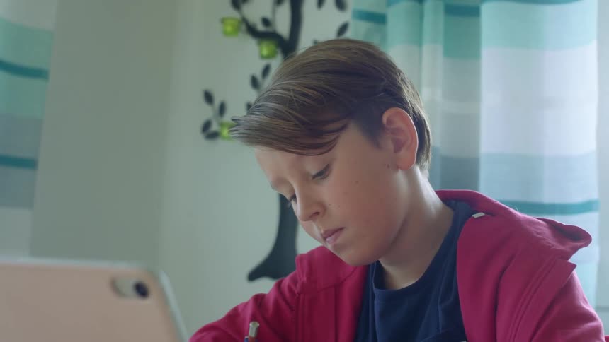 Boy wearing red hoodie sits near windows writing and viewing laptop screen