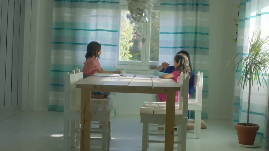 A woman helps her daughters with homework as they sit at their kitchen table