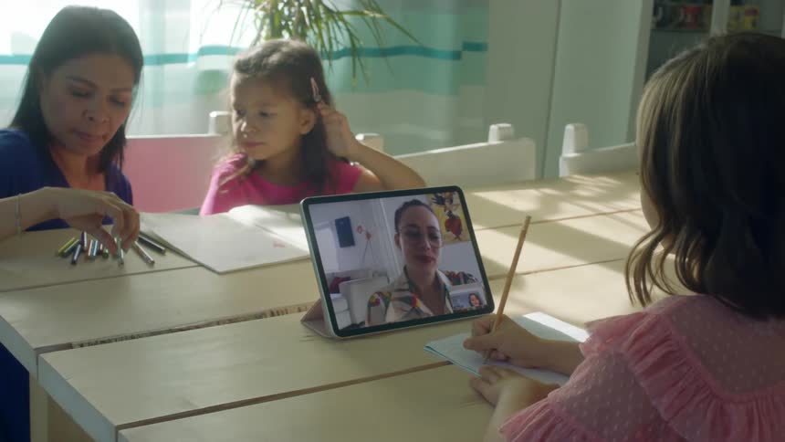 Mother helps one little girl with school work while another little girl works with a teacher on a tablet