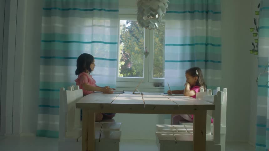 A pair of young girls do homework at a wooden table in a house during the day