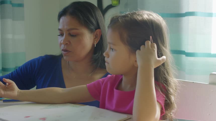 A young girl sitting at a table in house colors in a coloring book while her mother hands her colored pencils