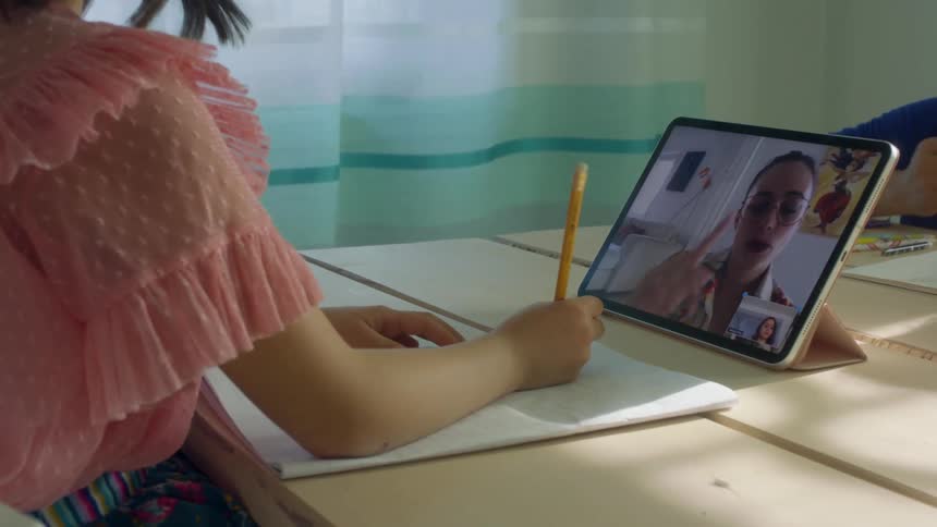 A girl sits at a table writing on paper as her laptop screen displays a person teaching a lesson inside during the day