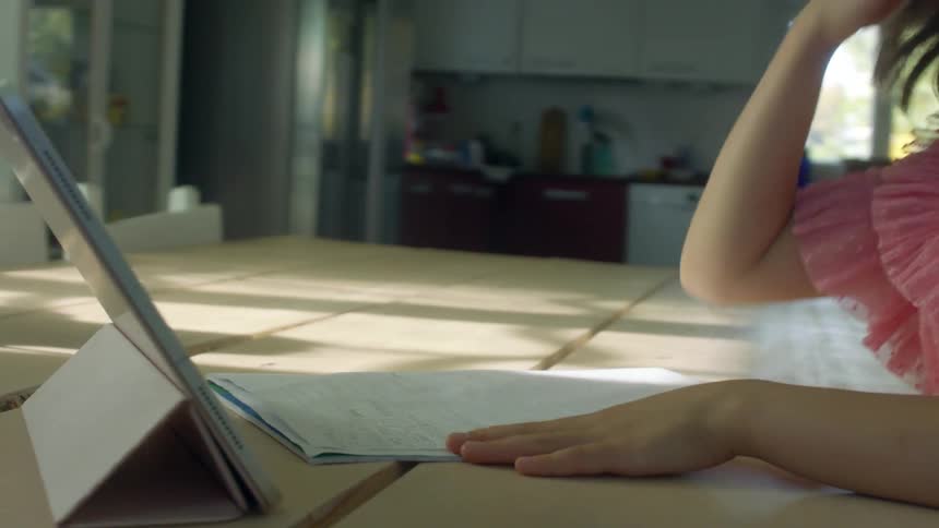 Three girls sit at a kitchen table doing homework as one girl looks at a tablet in front of her
