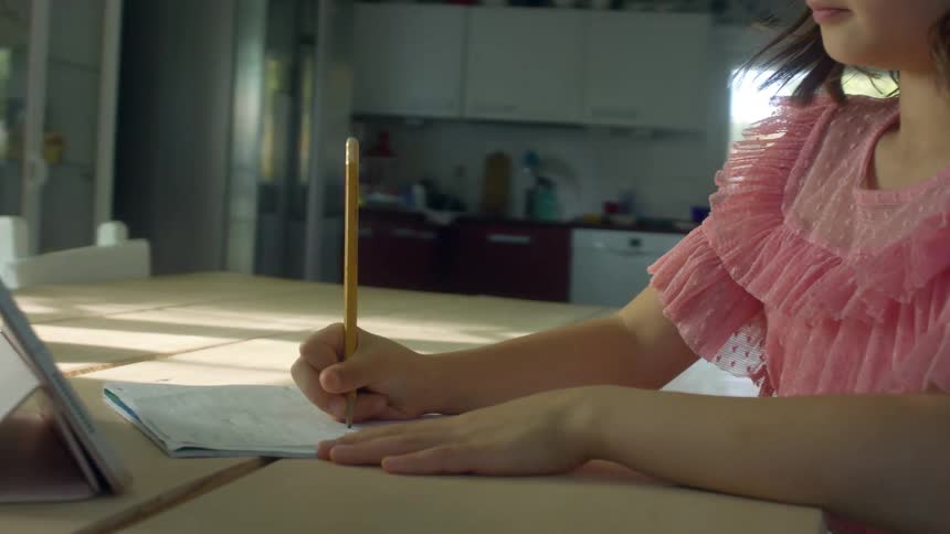 A young girl sits down at her kitchen table and watches her teacher on a tablet as her mother works with a younger sibling