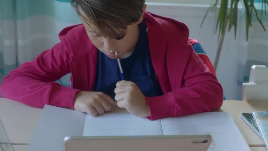 A young boy does his homework while sitting at a table indoors with a laptop in front of him during the day