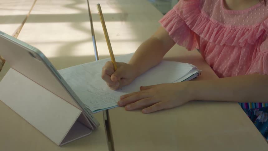 Girl with a pink blouse writes in a notebook next to a tablet
