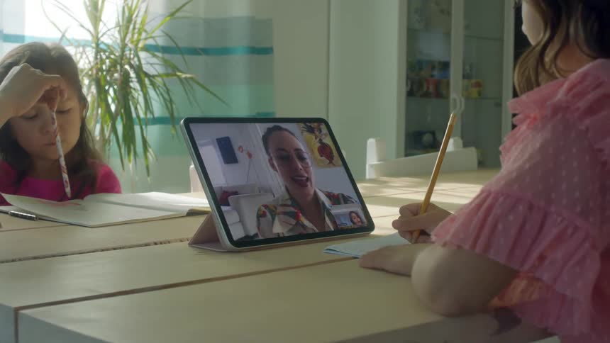 Two young girls work at a table indoors while one communicates with an adult via tablet