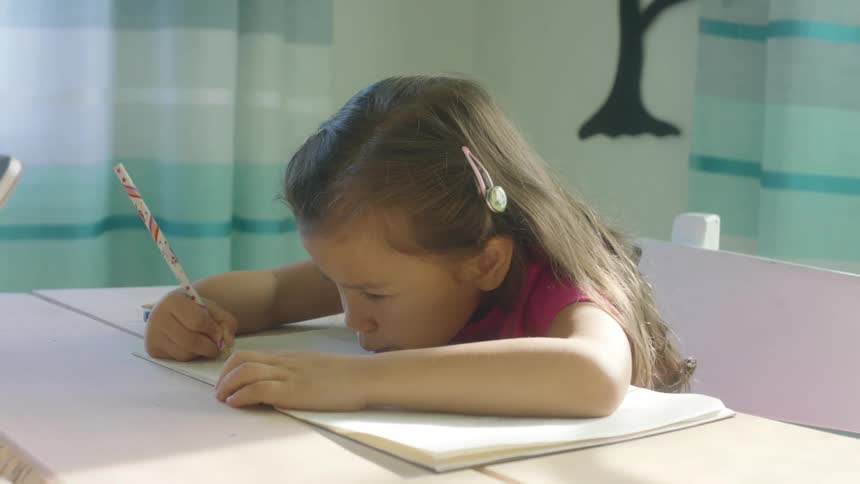 A young girls sits at a table writing on paper during the day at home