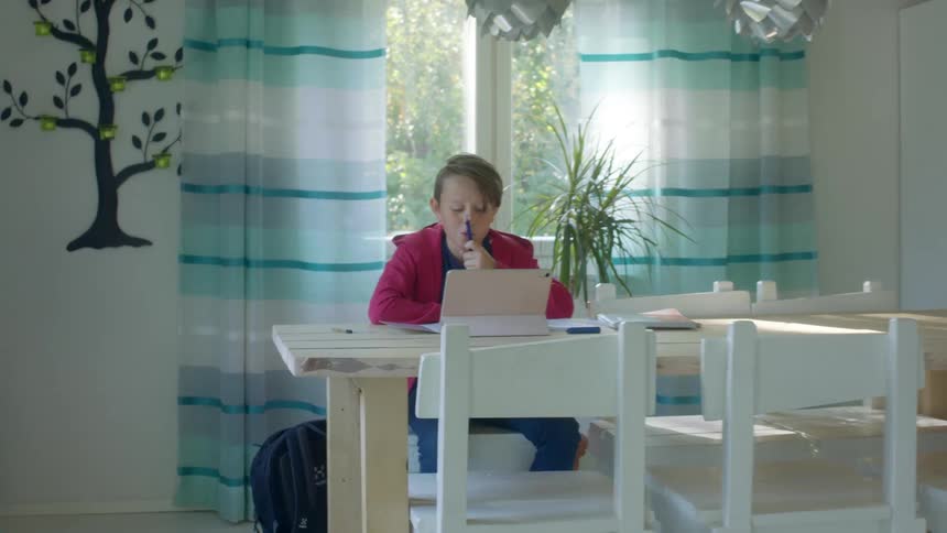 A boy sits at the dining room table doing homework with a laptop in front of him